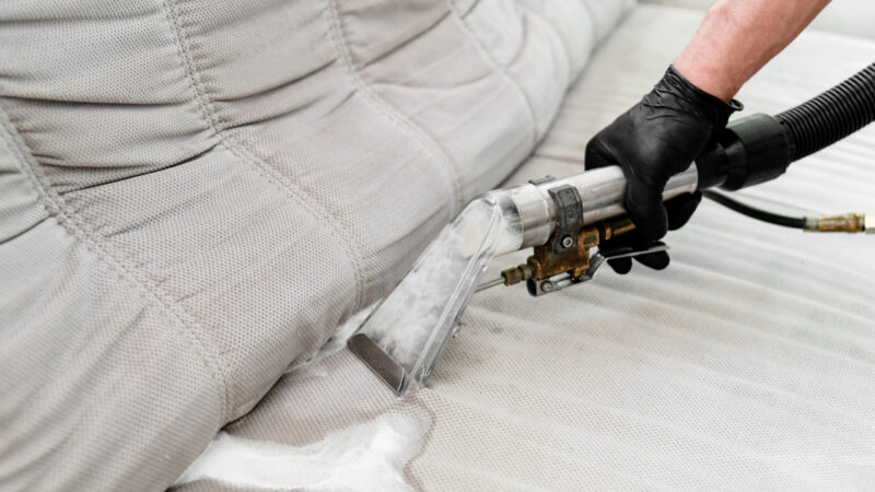 A technician is using specialized equipment to clean a fabric sofa in a living room. The process includes applying cleaning solution and extracting dirt.